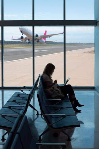 Vrouw achter haar laptop in de vertrekhal van een luchthaven, op de achtergrond stijgt een vliegtuig op